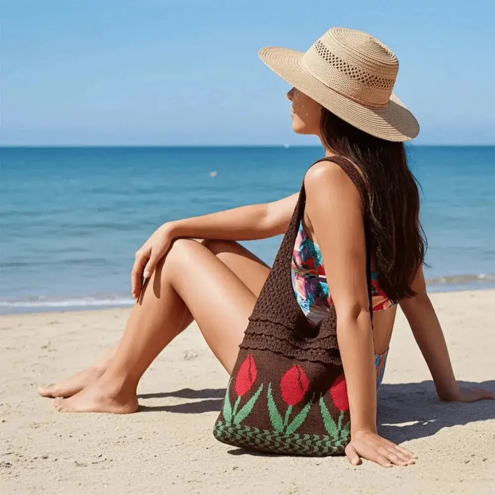 Woman in straw hat sitting on beach with a brown crochet tote bag featuring red tulip design.