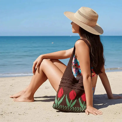 Woman in straw hat sitting on beach with a brown crochet tote bag featuring red tulip design.