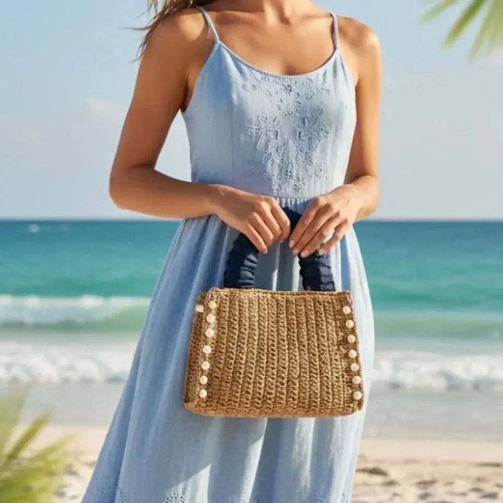 Woven raffia tote bag with ruffle handles and pearl accents, held by woman in blue dress at beach.