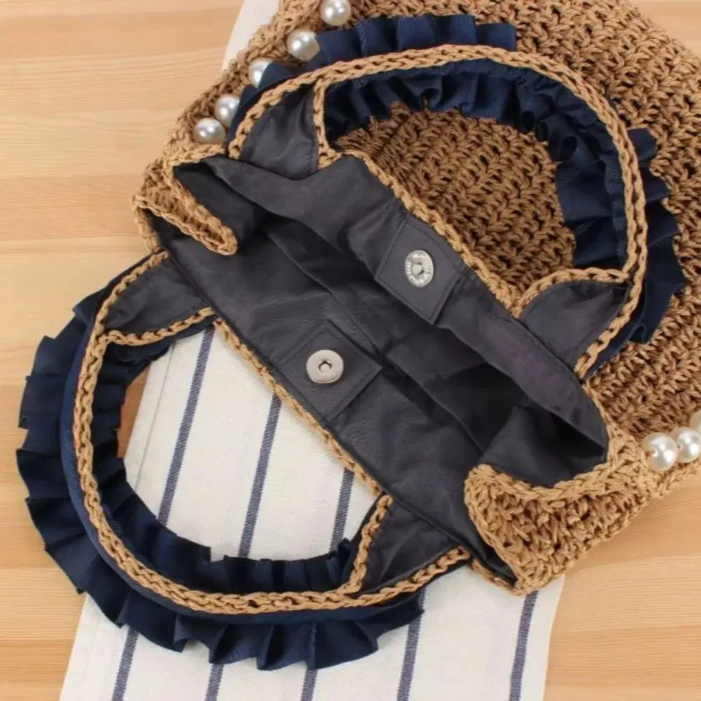Woven raffia tote bag with navy ruffle trim, pearl accents, and snap closure on wood surface.